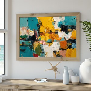 May include: Abstract art painting in a light wood frame. The artwork features bold brushstrokes of yellow, teal, white, orange, and pink. The painting is displayed on a wooden surface with decorative items.