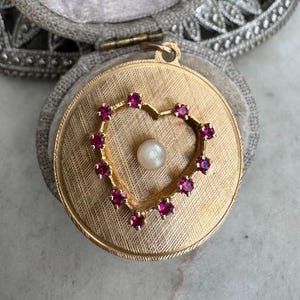 May include: A gold-toned locket with a heart-shaped ruby and pearl design. The locket is set in a gray velvet jewelry box. A gold flower pendant with a ruby center is visible in the background.