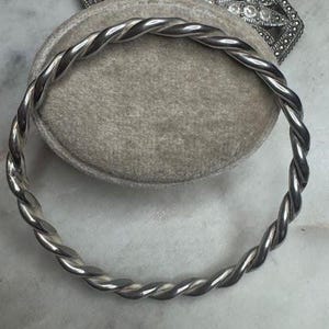 May include: A silver-toned, twisted rope bangle bracelet. The bracelet is circular and appears to be made of metal. The design features a rope-like twist throughout the entire bracelet. The bracelet is displayed on a gray, velvet-like surface.