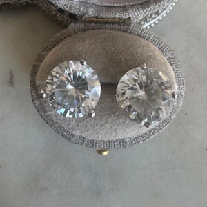 May include: A pair of sparkling, round-cut diamond stud earrings displayed in an open, gray velvet jewelry box. The earrings are set in a silver metal. The box is lined with a soft, light gray fabric.
