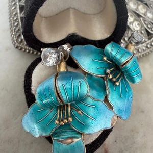 May include: A pair of earrings featuring bright turquoise enamel flower petals with gold-toned stamens and a single clear round-cut stone set at the top of each piece. The earrings are displayed inside a black heart-shaped velvet jewelry box.