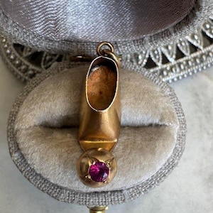 May include: A gold-coloured shoe charm with a round, pink gemstone at the base. The charm is set against a grey velvet background inside a decorative box. The shoe charm is a vintage piece.