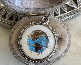 May include: A round pendant with a silver-colored frame, featuring a blue and brown globe design on a white background. The pendant is set in a gray velvet jewelry box. The globe design includes gold accents.