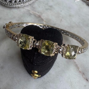 May include: A hinged bangle bracelet featuring three large, cushion-cut, pale yellow gemstones set in a silver-toned metal frame. The band displays an intricate, textured pattern along its length, with small, clear, sparkling accents surrounding each stone.