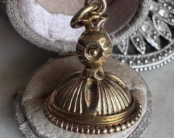May include: A gold-colored antique wax seal with a decorative design, resting inside a gray velvet-lined case. The seal has a detailed, ornate pattern and a ring at the top. The case is open, revealing the seal and its soft interior.