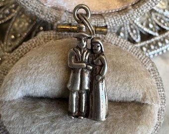 May include: A small, silver-toned charm depicting a man and woman in traditional attire. The charm is suspended from a small ring and is set against a soft, gray background. The figures are detailed, with visible clothing and facial features.