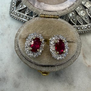 May include: A pair of oval-cut red gemstone stud earrings, each surrounded by a halo of small clear stones. The earrings are set in yellow metal and are displayed inside a beige, oval-shaped, fabric-lined jewelry box with a hinged lid.