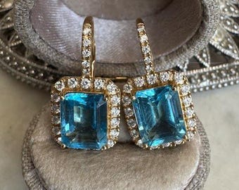 May include: A pair of gold-toned earrings with rectangular, light blue gemstones. Each earring is surrounded by a halo of small, clear gemstones. The earrings are displayed in a gray, velvet-lined jewelry box.