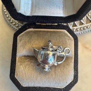 May include: A miniature silver teapot charm displayed in a black velvet jewellery box with a beige interior. The teapot has a handle, spout, and lid. The box is open, revealing the charm. A decorative item is visible in the background.