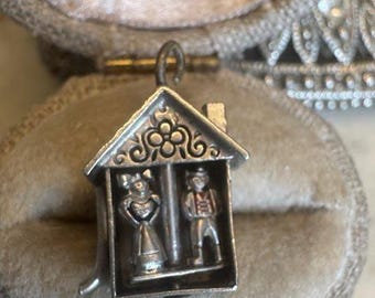 May include: A silver charm in the shape of a house, featuring two small figures inside. The charm has a decorative roof and is set against a soft, neutral background, suggesting a vintage or antique piece.