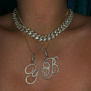 May include: A gold chain necklace with a diamond-encrusted cuban link chain and two pendant necklaces. The pendants are diamond-studded, cursive letters "Y" and "B". The jewelry is displayed on a person's chest.