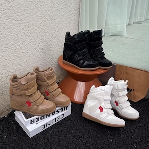 May include: Four pairs of wedge sneakers in various colors are displayed. There are black, tan, and white pairs, each with multiple hook-and-loop straps. The tan sneakers have a red accent. The shoes are on a small orange table and a stack of books.