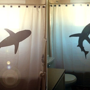 May include: Two white shower curtains with a black shark silhouette printed on each. The shark on the left is swimming horizontally, while the shark on the right is swimming vertically.