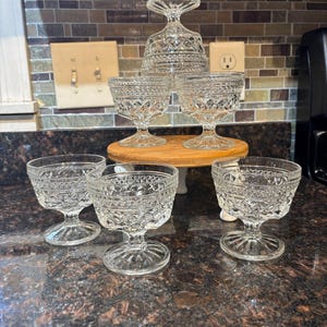 May include: A collection of clear, vintage glass dessert dishes. Six stemmed glasses with a diamond pattern are displayed on a wooden cake stand and countertop. One glass is upside down. The glasses are ideal for serving desserts.