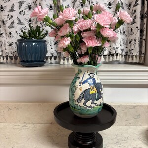 May include: A white ceramic vase with a painted scene of a person riding a horse. The vase is filled with pink carnations and sits on a black pedestal.
