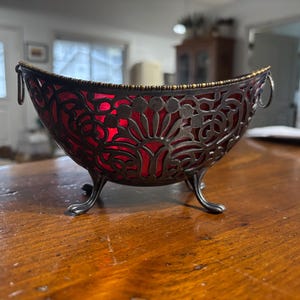 May include: A red glass bowl with a silver filigree design and three legs. The bowl has a gold rim and is sitting on a wooden table.