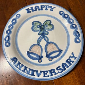 May include: A white ceramic plate with blue and green designs. The plate has the words "Happy Anniversary" around the edge and two bells with a bow in the center.