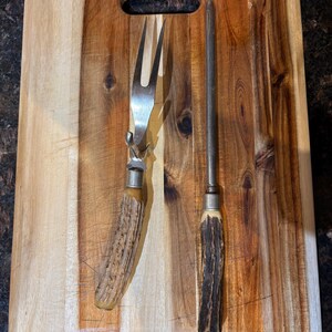 May include: Two vintage kitchen utensils with antler handles on a wooden cutting board. The utensils are a carving fork and a knife.