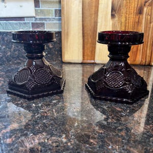 May include: Two dark red glass candle holders with a decorative design. The holders have a round base and a wide, flat top.