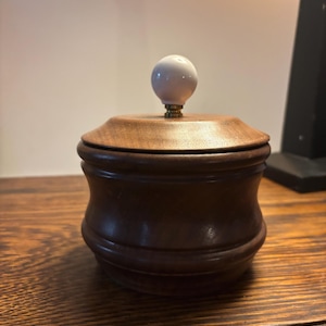 May include: A small wooden box with a white ceramic knob on the lid. The box is made of dark wood and has a rounded shape.