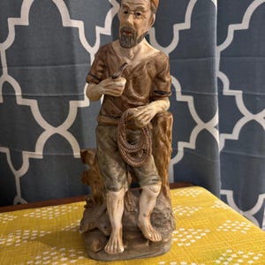 May include: A porcelain figurine of a man with a brown hat, beard, and brown shirt holding a rope and a pipe. He is standing on a base with a tree trunk behind him.