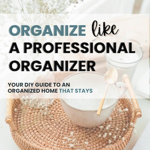 May include: A round woven rattan tray holds a large mug with a golden spoon, a glass, and a wooden rolling pin. The text "ORGANIZE like A PROFESSIONAL ORGANIZER" is displayed above the tray, with the text "YOUR DIY GUIDE TO AN ORGANIZED HOME THAT STAYS" below.