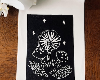 May include: A black and white linocut print featuring three stylized mushrooms with detailed caps and stems, set against a dark background with diamond-shaped accents. The print is on white paper, with fern-like foliage at the base.