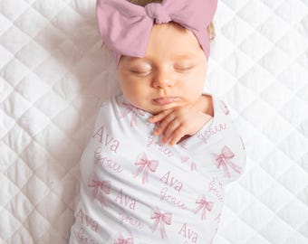 May include: A newborn baby wrapped in a white swaddle blanket and wearing a matching pink bow headband. The swaddle is patterned with pink bows and the names "Ava" and "Grace". The baby is lying on a white quilted surface.