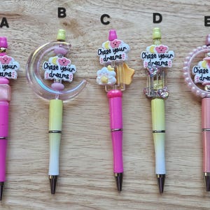 May include: Five pink and yellow pens with different charms. The charms include a crescent moon, a cloud, a cage, and a circle. Each charm has the text "Chase your dreams" on it.