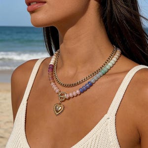 May include: A layered necklace featuring a gold-tone chain and a strand of multi-colored beads in shades of pink, blue, and green. The beaded strand includes a gold-tone toggle clasp and a dangling gold-tone heart-shaped pendant with a textured border.