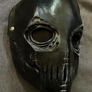 Puede incluir: Una m&aacute;scara facial completa de color negro con cuencas oculares profundas y malla oscura, una superficie texturizada con detalles de costuras simuladas en las mejillas y la barbilla, y un remache met&aacute;lico plateado en el lateral.