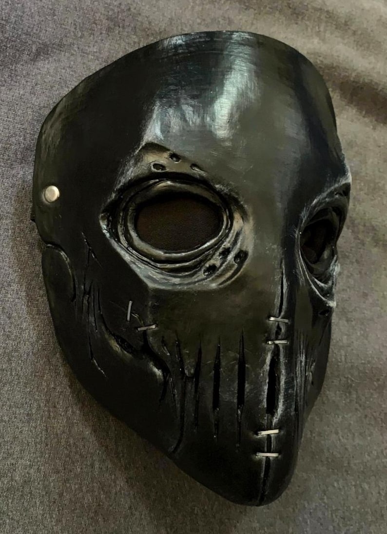Puede incluir: Una m&aacute;scara facial completa de color negro con cuencas oculares profundas y malla oscura, una superficie texturizada con detalles de costuras simuladas en las mejillas y la barbilla, y un remache met&aacute;lico plateado en el lateral.