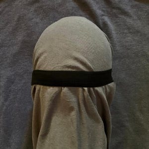 Puede incluir: Un cubrecabezas de tela gris claro liso con una parte superior redondeada y suave, y una banda el&aacute;stica negra horizontal que rodea el centro, creando pliegues suaves en el material debajo de la banda para un aspecto drapeado.