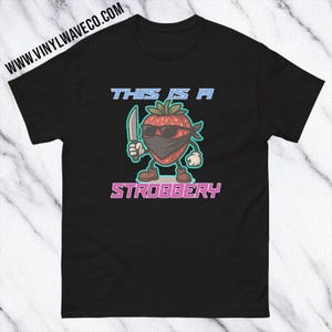 May include: Black t-shirt featuring a cartoon strawberry character wearing sunglasses and a bandana, holding a knife. The text "THIS IS A STROBBERY" is displayed in blue and pink. The website address "WWW.VINYLWAVECO.COM" is printed at the top.