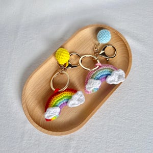 May include: Two handmade crochet rainbow keychains with gold-colored hardware. One rainbow is red, orange, yellow, green, blue, and purple with white clouds. The other is pink, blue, and purple with white clouds. Each keychain has a small ball at the top.