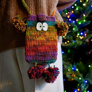 May include: A colorful crocheted crossbody bag with pom-poms and googly eyes. The bag is made with a rainbow of colors, including red, orange, yellow, green, blue, purple, and brown.