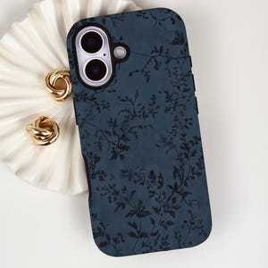 May include: A navy blue phone case with a black floral pattern rests beside a white, scalloped dish holding two gold-colored knot earrings. The phone case has a dual-lens camera cutout.