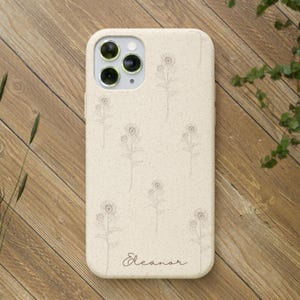 May include: A beige phone case with a floral design and the name "Eleanor" in script. The case has a speckled texture and a camera cutout. The floral pattern consists of delicate line drawings of flowers.