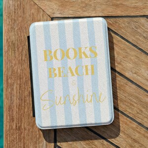 May include: A rectangular e-reader case featuring a light blue and white vertical striped pattern. The text "BOOKS BEACH & Sunshine" is printed in gold lettering on the front cover. The case has a black spine and a textured surface finish.