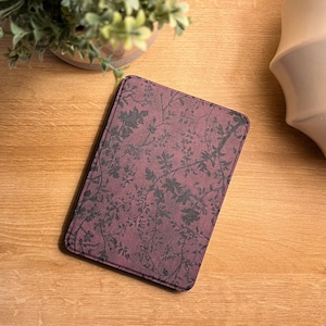 May include: A rectangular, burgundy-colored item with a floral pattern in dark gray. The item is on a wooden surface, next to a small plant and a white, decorative vase. The floral design features leaves and branches.