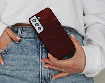 May include: A burgundy phone case with a flowing, abstract design. The case has a camera cutout with three lenses and a flash. The phone case is held in a person's hand, with the person wearing a white sweatshirt and blue jeans.