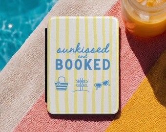 May include: A rectangular e-reader case featuring a white background with vertical yellow stripes. The text "sunkissed and BOOKED" is printed in blue, accompanied by blue line-art icons of a beach tote bag, a beach sign, and sunglasses.