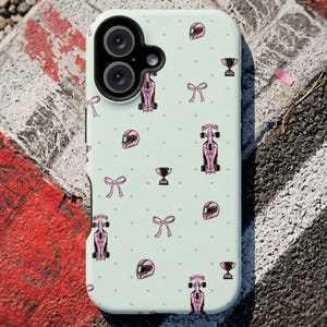 May include: A light green phone case featuring a pattern of pink race cars, helmets, bows, and trophies. The case has a dual-lens camera cutout and is set against a backdrop of a red and white painted surface and asphalt.