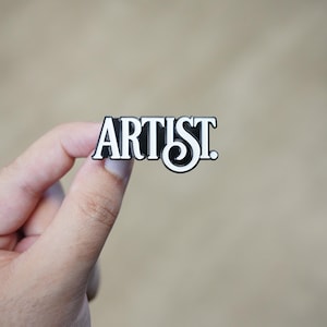 May include: A black and white enamel pin with the word "ARTIST" in a bold, stylized font. The pin features a black background with white lettering and a white border. The pin is held in a hand against a neutral background.