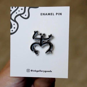 Puede incluir: Pin de esmalte negro con un diseño de figura estilizada. El pin está sobre una tarjeta blanca con el texto "ENAMEL PIN" y el identificador de Instagram "@inkgallerygoods". El pin tiene un borde plateado.