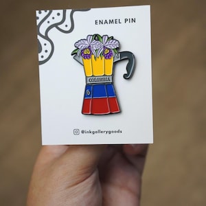 May include: Enamel pin featuring a coffee maker design with the colors of the Colombian flag: yellow, blue, and red. The coffee maker is topped with purple and green flowers. The pin is on a white card that reads "ENAMEL PIN" and has the Instagram handle "@inkgallerygoods".