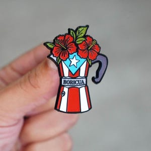 May include: A colorful enamel pin in the shape of a coffee pot, featuring the Puerto Rican flag design. The pin is adorned with red hibiscus flowers and the word "BORICUA".