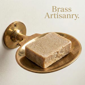 Pode incluir: Um saboneteira de latão com uma barra de sabão. A saboneteira é montada na parede e tem um design clássico. O sabão é de cor castanho claro e fica no centro da saboneteira. O texto "Brass Artisanry" está no canto superior direito.