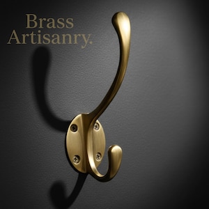 Puede incluir: Un gancho de pared de latón con un diseño curvo, con una parte superior redondeada y un gancho inferior. El gancho está montado sobre una base ovalada con cuatro cabezas de tornillo. El texto "Brass Artisanry" está en la esquina superior izquierda.