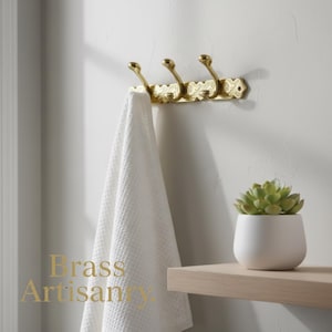 May include: A gold-colored, three-hook wall-mounted rack with ornate detailing, holding a white waffle-weave towel. A small white pot with a green succulent sits on a wooden shelf. The text "Brass Artisanry" is visible.